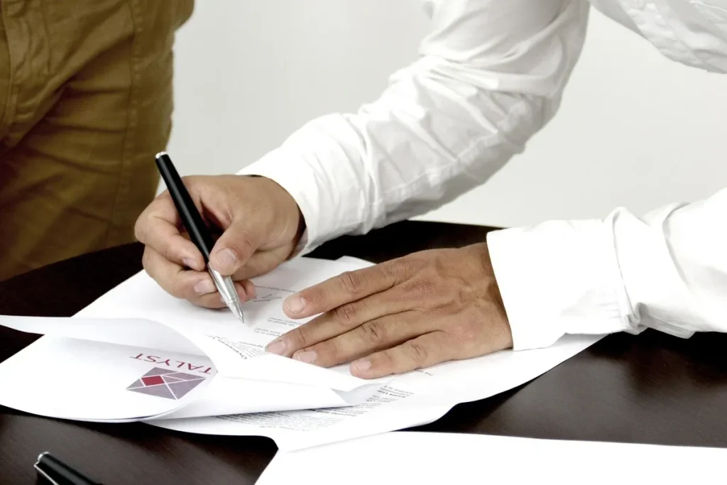 Homem assinando documentos de carta de crédito em uma mesa, com outro indivíduo ao lado.