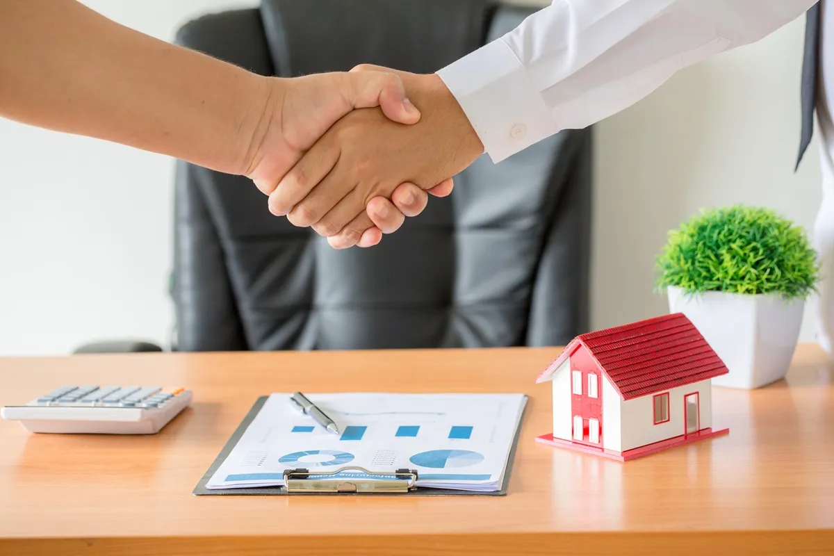 Duas pessoas apertam as mãos em um escritório após a negociação de um imóvel, representado por uma miniatura de casa sobre a mesa.