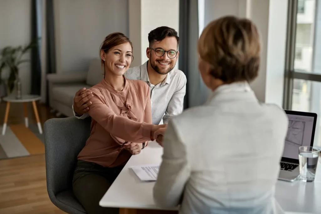 Casal sorridente aperta a mão de uma consultora financeira durante reunião, ilustrando a satisfação ao fechar um acordo, possivelmente relacionado a uma carta de crédito sem anuidade.