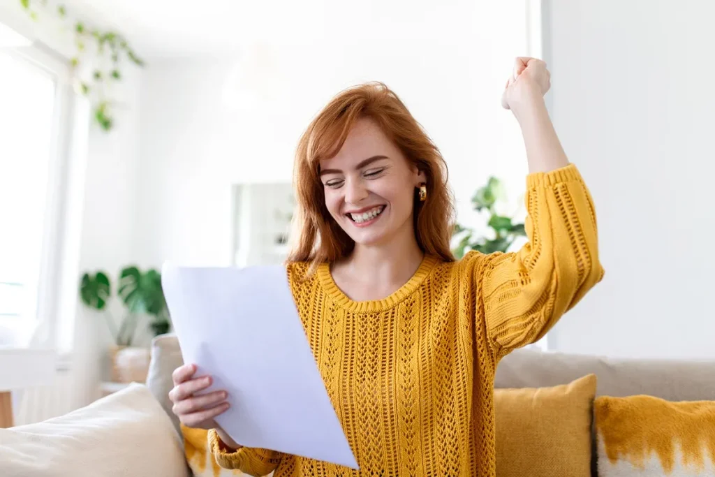 Mulher sorridente com suéter amarelo comemora ao ler um documento, sugerindo aprovação de uma carta de crédito sem anuidade, financiamento ou empréstimo.