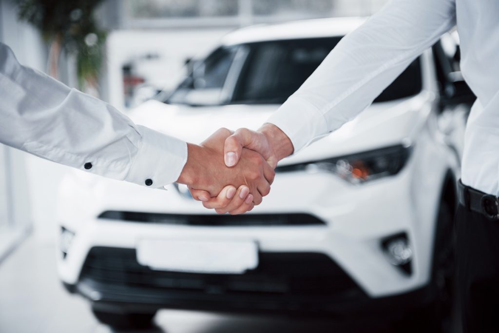 Duas pessoas apertando as mãos em um acordo de compra de veículo, com um carro branco ao fundo, simbolizando consórcio ou financiamento automotivo.