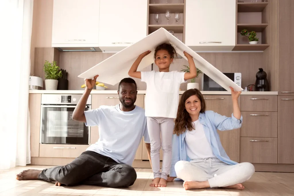 Família feliz (pai, mãe e filha pequena) em uma cozinha moderna, com a criança segurando um "telhado" de papel acima de suas cabeças, simbolizando a conquista da casa própria através do consórcio de imóveis.