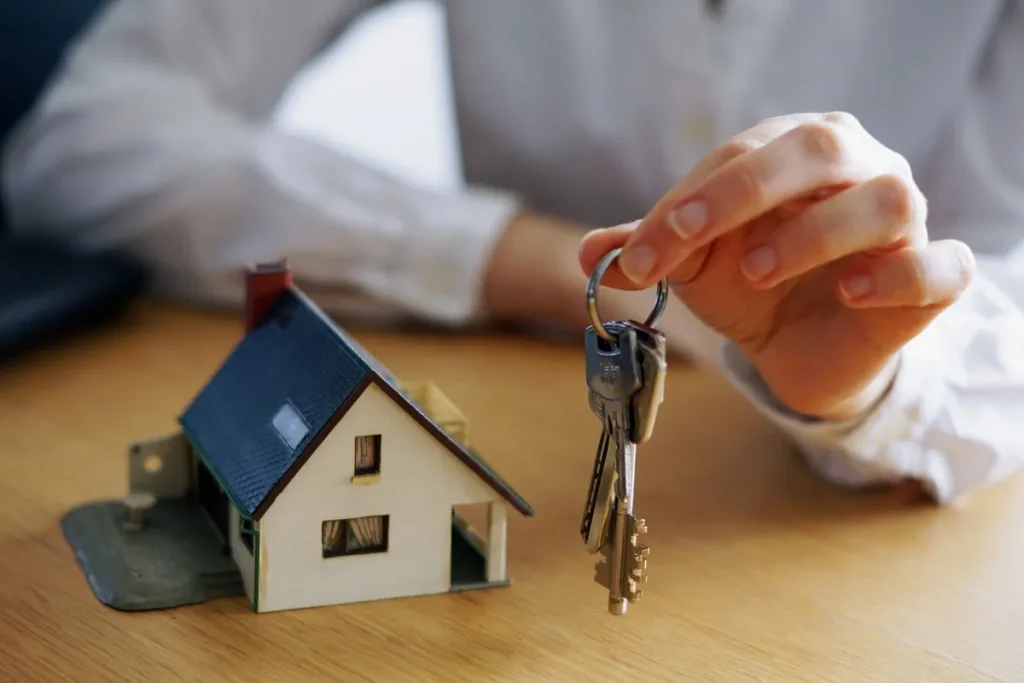 Mão segurando um chaveiro com várias chaves ao lado de uma miniatura de casa sobre uma mesa de madeira, representando a compra de imóveis por meio de consórcio.