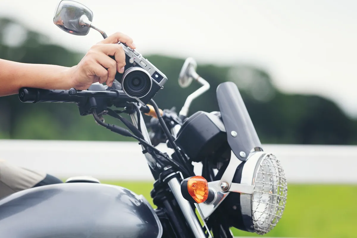 Close-up de uma motocicleta clássica com um viajante segurando uma câmera fotográfica, capturando momentos durante uma viagem.