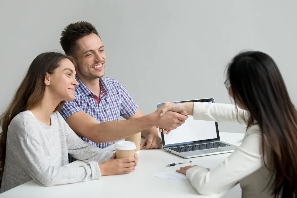 Casal sorridente apertando a mão de uma consultora durante uma reunião, simbolizando a conclusão de um acordo financeiro ou de consórcio.