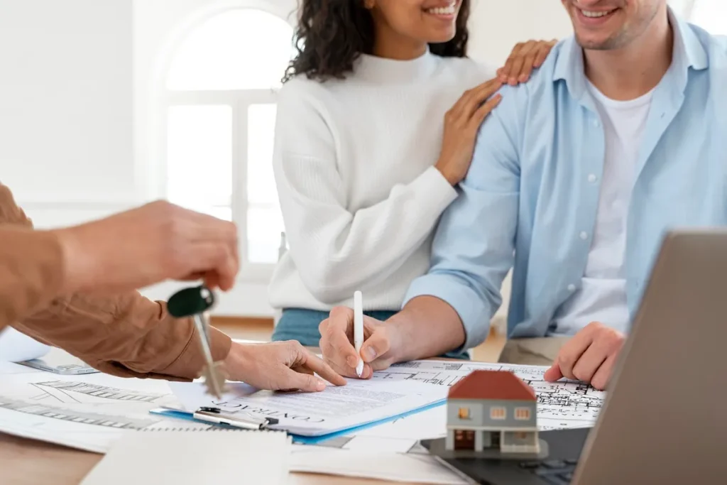 Casal assinando contrato de consórcio imobiliário enquanto recebe chaves, simbolizando a aquisição de um imóvel. Miniatura de casa e laptop sobre a mesa reforçam o conceito de planejamento financeiro e investimento em bens.