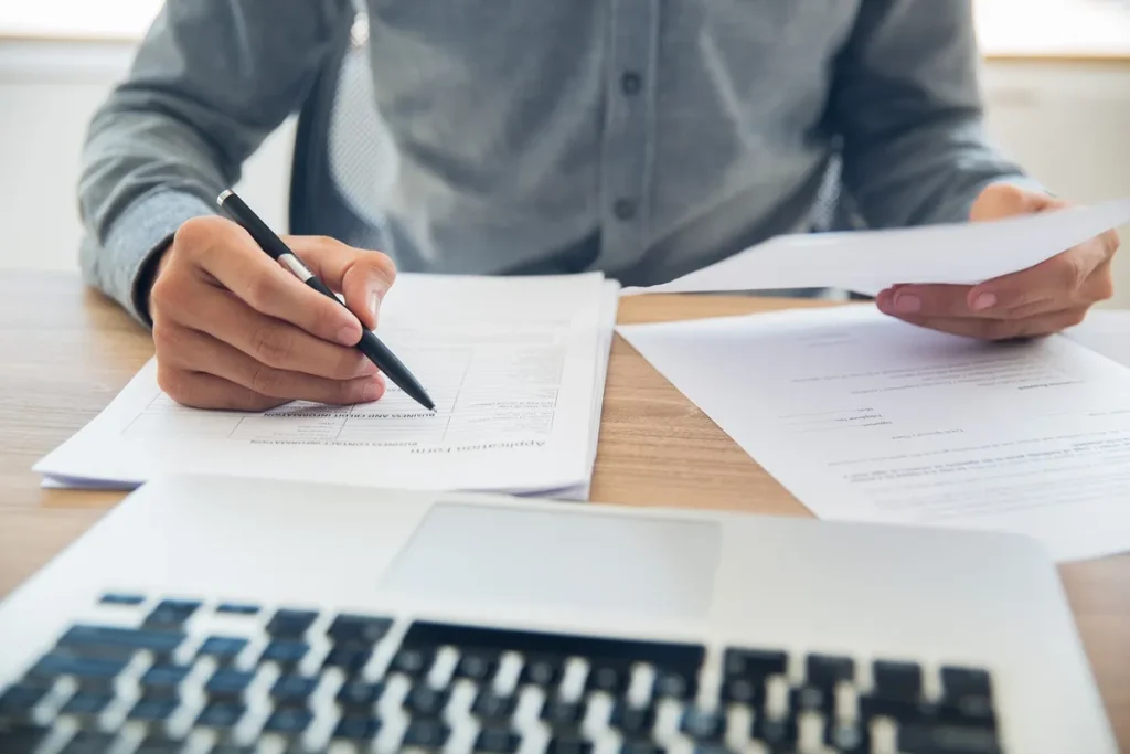 Pessoa analisando e preenchendo documentos financeiros sobre uma mesa, com um laptop aberto ao fundo, representando a gestão de crédito e financiamentos.