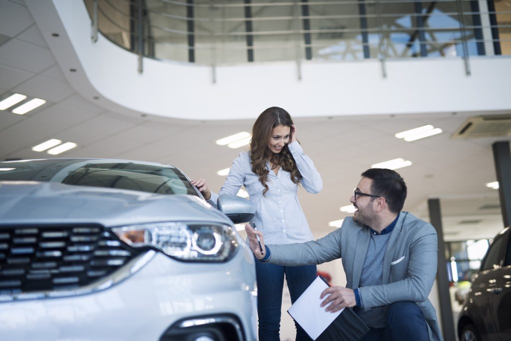 Mulher sorridente analisa um carro em uma concessionária enquanto conversa com um consultor que explica detalhes sobre consórcio automotivo.