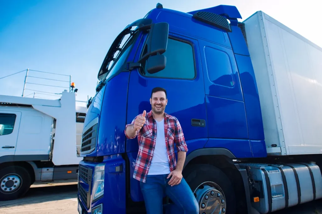 Caminhoneiro sorridente vestindo camisa xadrez faz sinal de positivo ao lado de um caminhão azul, com outro veículo ao fundo.