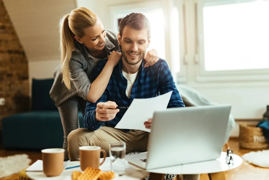 Casal feliz analisando documentos financeiros em casa, com laptop e café sobre a mesa, simbolizando planejamento para aquisição de imóvel ou investimento.