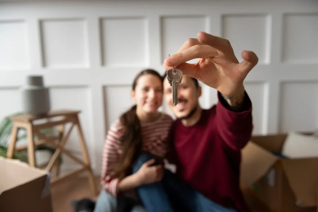 Casal sorridente segura as chaves da nova casa, cercado por caixas de mudança e decoração aconchegante.