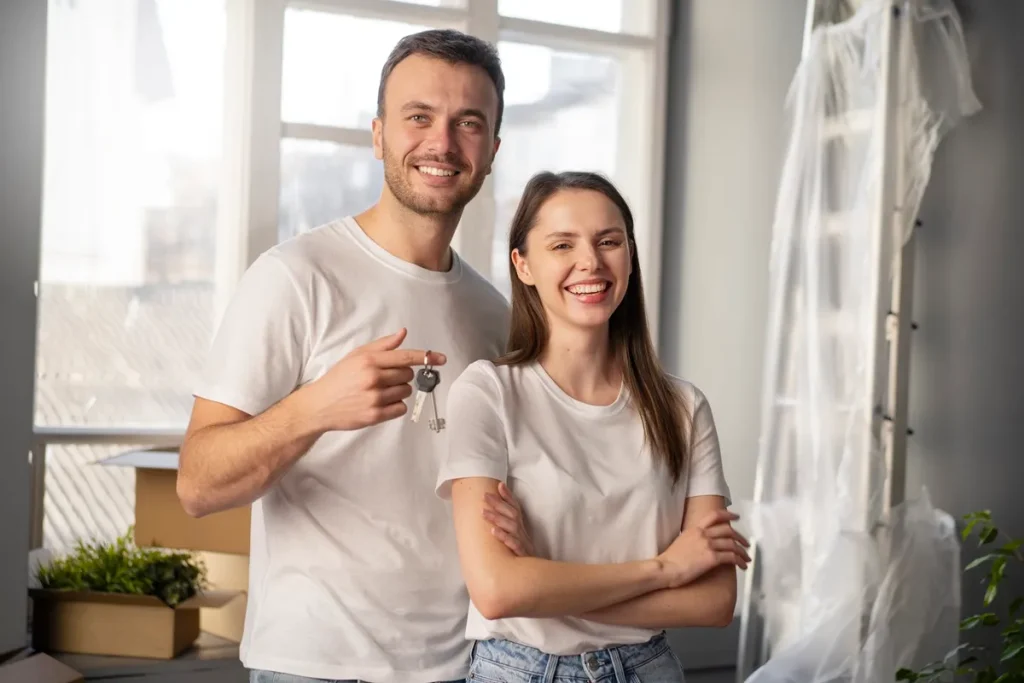 Casal sorridente segura as chaves da casa nova, rodeado por caixas de mudança e decoração moderna.