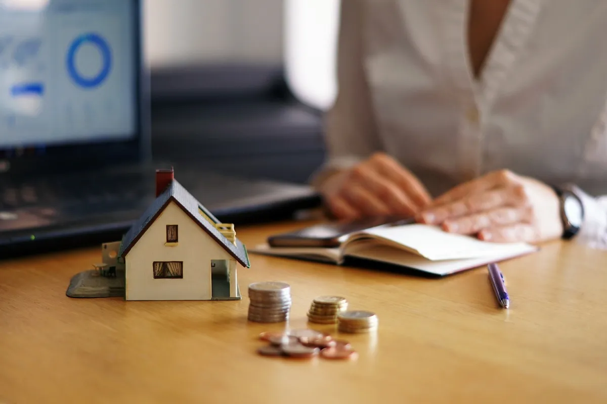 Miniatura de casa ao lado de pilhas de moedas em uma mesa, simbolizando investimento imobiliário, enquanto uma pessoa escreve em um caderno com um notebook ao fundo.