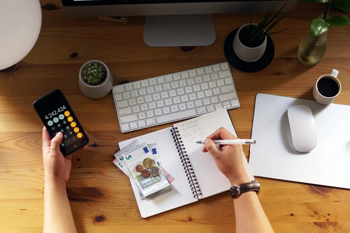 Pessoa organizando finanças pessoais em um ambiente de home office, segurando um celular com calculadora aberta e anotando valores em um caderno, com dinheiro em espécie ao lado.