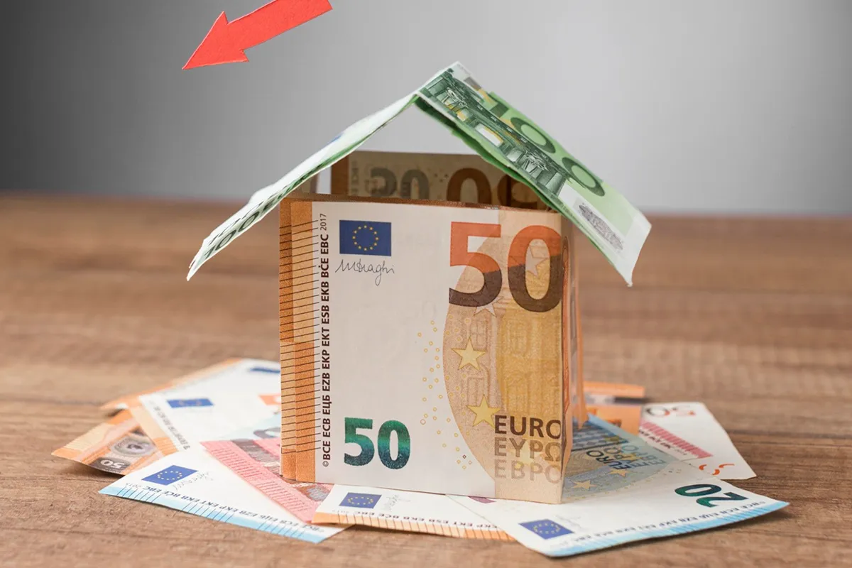 Notas de euro formando uma casa em miniatura com seta vermelha apontando para baixo, simbolizando consórcio, economia ou queda de preços no setor imobiliário.