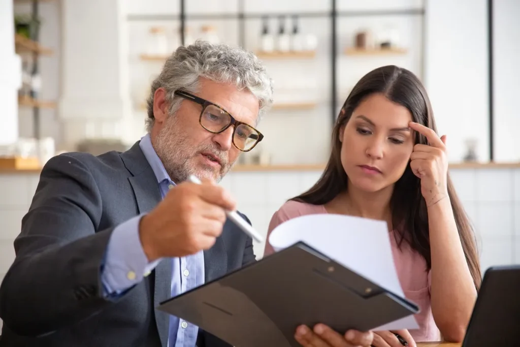 Homem mais velho, de terno e óculos, orienta uma mulher jovem com expressão séria enquanto analisa documentos, representando atendimento sobre consórcio ou planejamento financeiro.