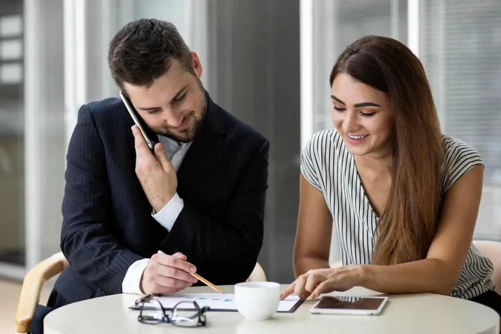 Homem de terno e mulher casual analisando documentos financeiros em uma reunião, enquanto conversam ao telefone e usam tablet