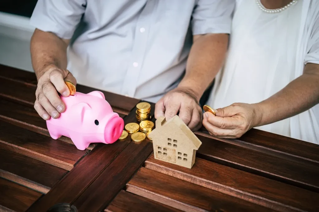 Mãos de um casal colocando moedas em cofre de porquinho ao lado de uma miniatura de casa e pilhas de moedas sobre a mesa, simbolizando economia e consórcio imobiliário.