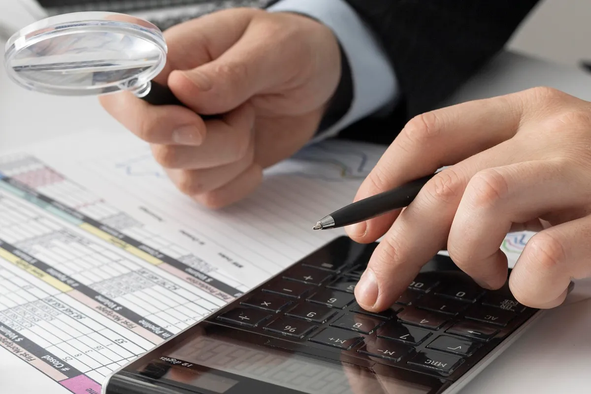 Pessoa usando uma calculadora enquanto segura uma lupa sobre uma planilha com colunas e valores coloridos, analisando custos administrativos.