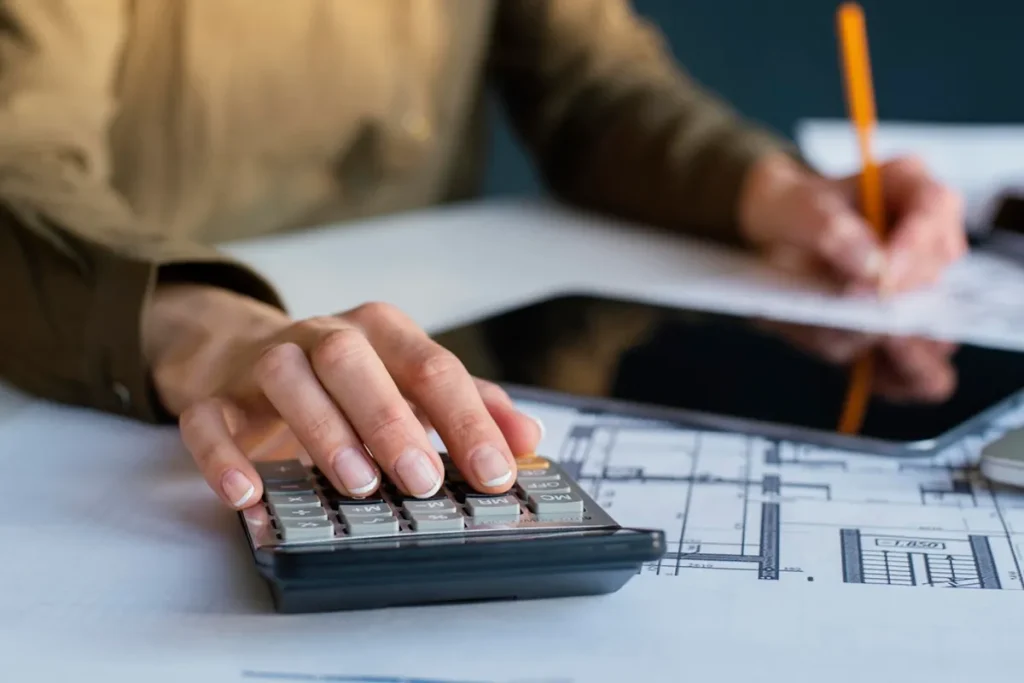Pessoa utilizando uma calculadora sobre uma planta arquitetônica enquanto faz anotações com lápis, representando o planejamento financeiro de uma construção ou reforma.