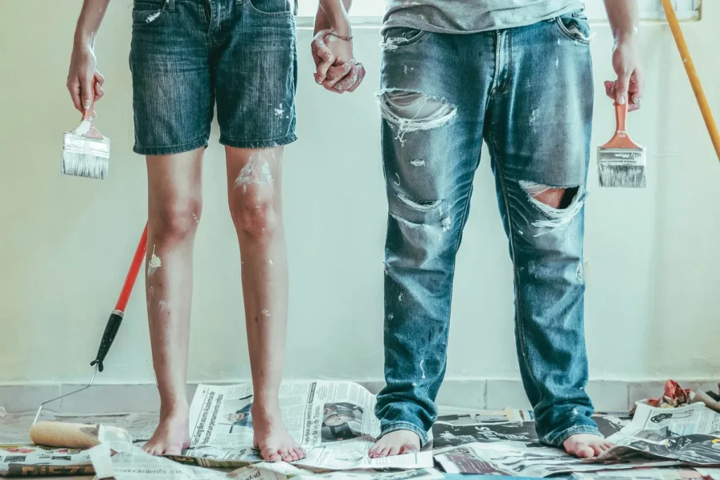 Casal segurando pincéis e de mãos dadas durante reforma caseira, com roupas manchadas de tinta sobre jornal no chão