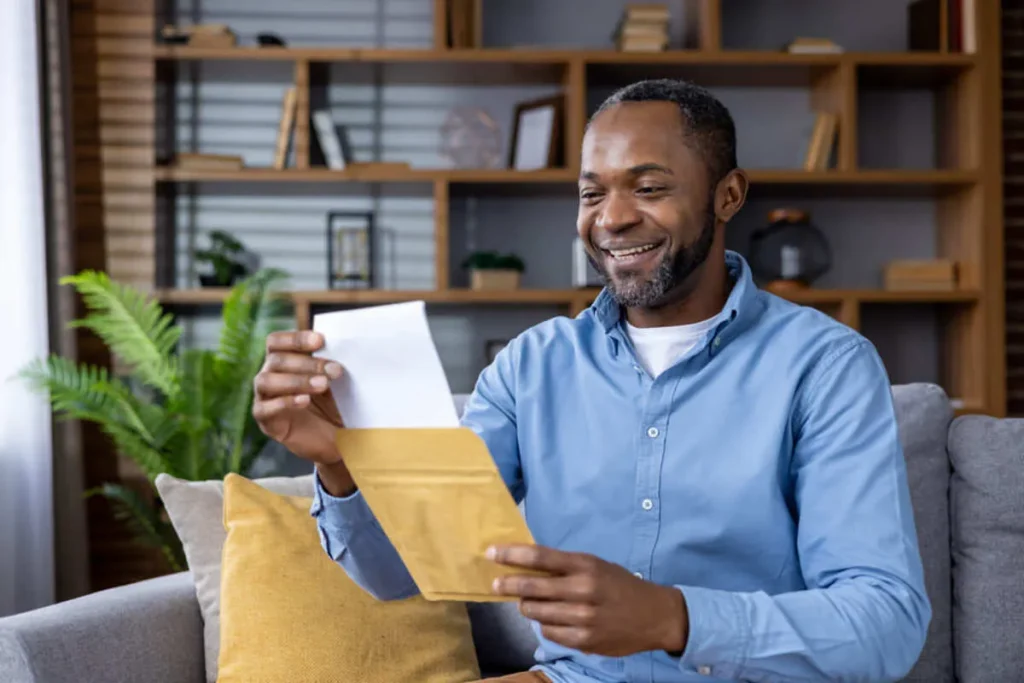 Homem negro de meia-idade sorrindo e abrindo um envelope em sua sala de estar.