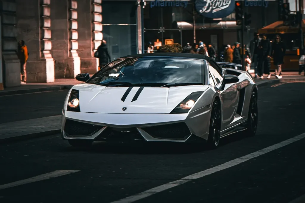 Um carro de luxo conversível, modelo Lamborghini Gallardo, branco com listras pretas, em movimento em uma rua urbana.