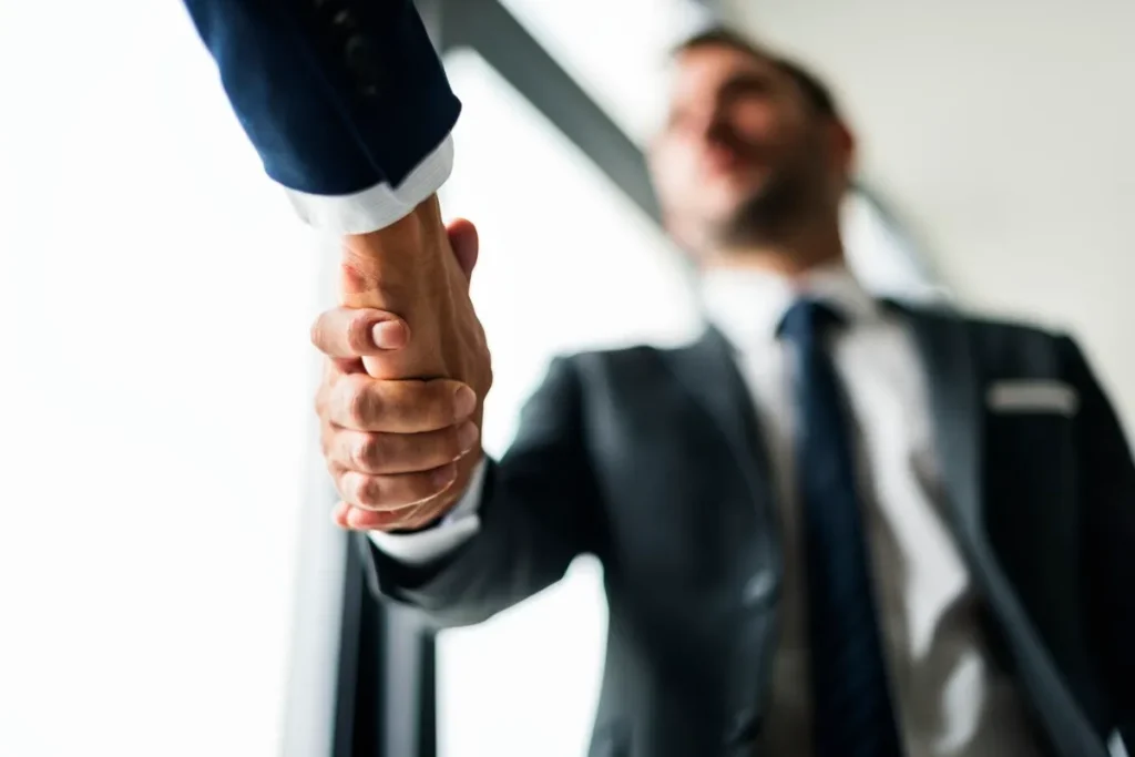 Aperto de mão entre dois homens de terno em ambiente corporativo, simbolizando um acordo comercial ou parceria.
