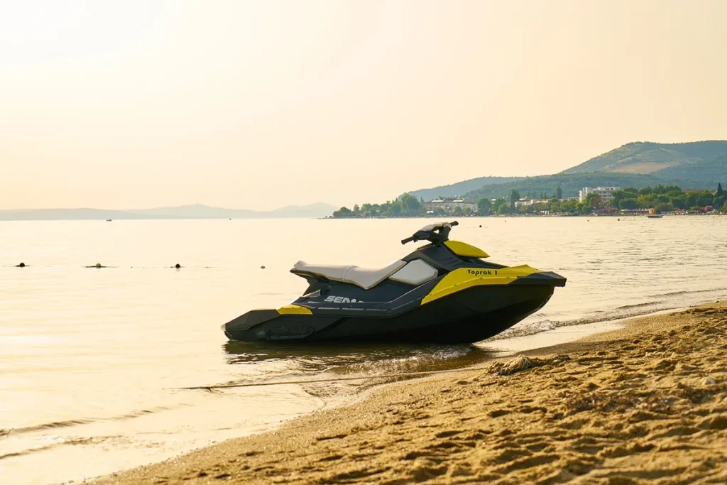 Jet ski amarelo e preto na areia da praia, com vista para o mar e montanhas ao fundo.