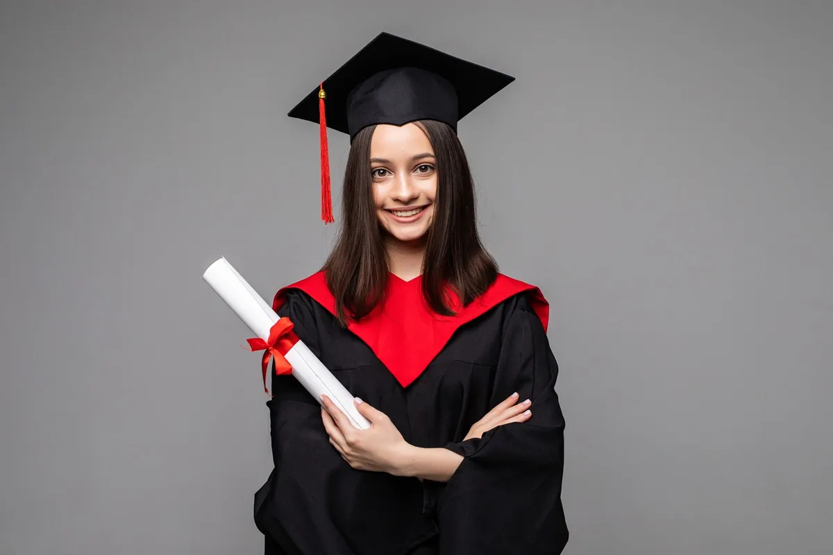Imagem de uma jovem sorridente vestindo capelo e beca de formatura, segurando um diploma com fita vermelha.