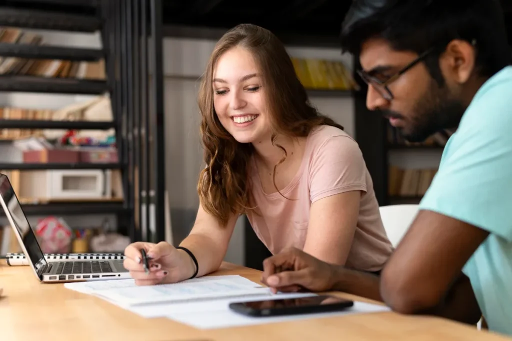 Dois jovens estudando juntos, analisando documentos em uma mesa, com laptop e celular ao fundo