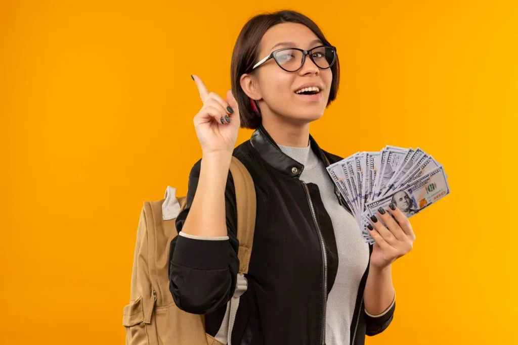 Mulher com óculos segurando notas de 100 dólares, sorrindo e apontando para cima com fundo amarelo