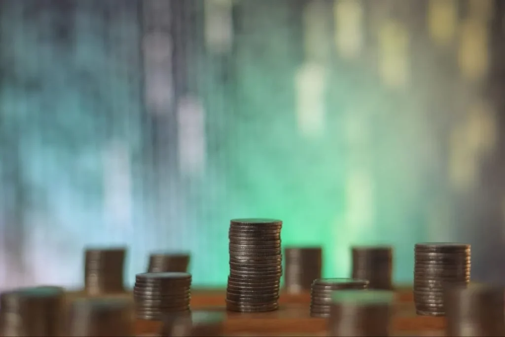 Close-up de moedas empilhadas em uma mesa, representando a acumulação de dinheiro e o fundo de reserva, com um fundo desfocado de gráficos financeiros em cores verde e azul.