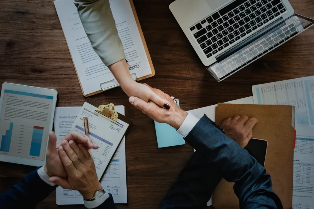 Aperto de mão profissional entre dois executivos em ambiente corporativo, com documentos e dispositivos eletrônicos sobre a mesa.
