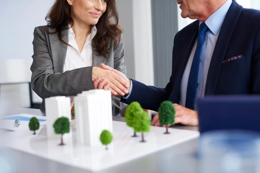 Dois profissionais de negócios, um homem e uma mulher, realizando um aperto de mão em frente a uma maquete de um prédio, representando um acordo de compra ou investimento imobiliário.