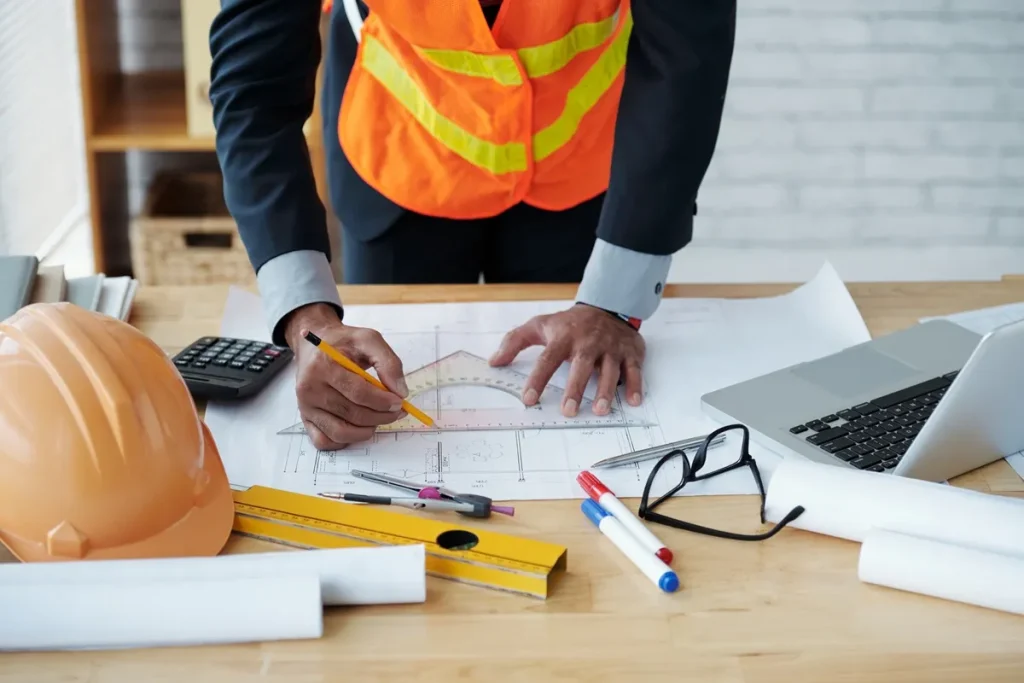 Profissional de construção civil analisando projetos arquitetônicos, utilizando régua, esquadros e lápis sobre uma mesa com calculadora, laptop e material de escritório.