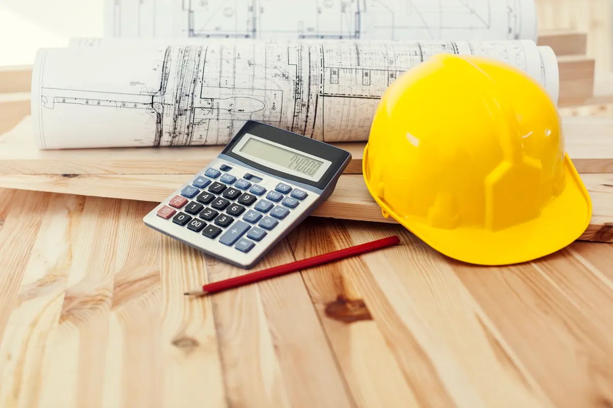Calculadora, capacete de segurança e plantas arquitetônicas sobre uma mesa de madeira, elementos essenciais no planejamento e execução de projetos de construção.