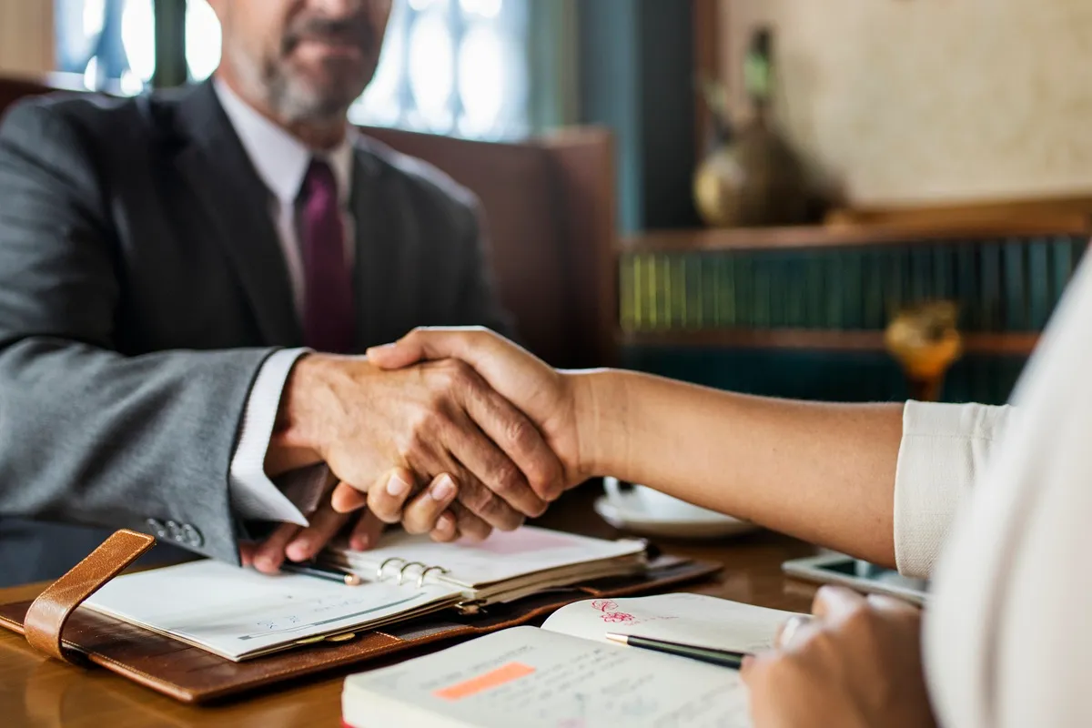 Aperto de mãos entre duas pessoas em reunião de negócios com anotações e agenda sobre a mesa.