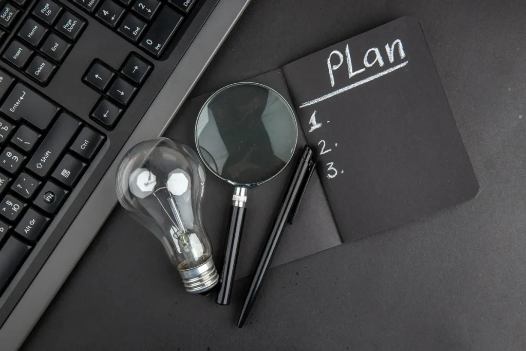 Caderno preto com a palavra “Plan” e lista enumerada ao lado de teclado, lâmpada, lupa e caneta sobre mesa escura.