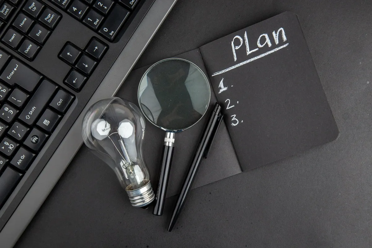 Caderno preto com a palavra “Plan” e lista enumerada ao lado de teclado, lâmpada, lupa e caneta sobre mesa escura.