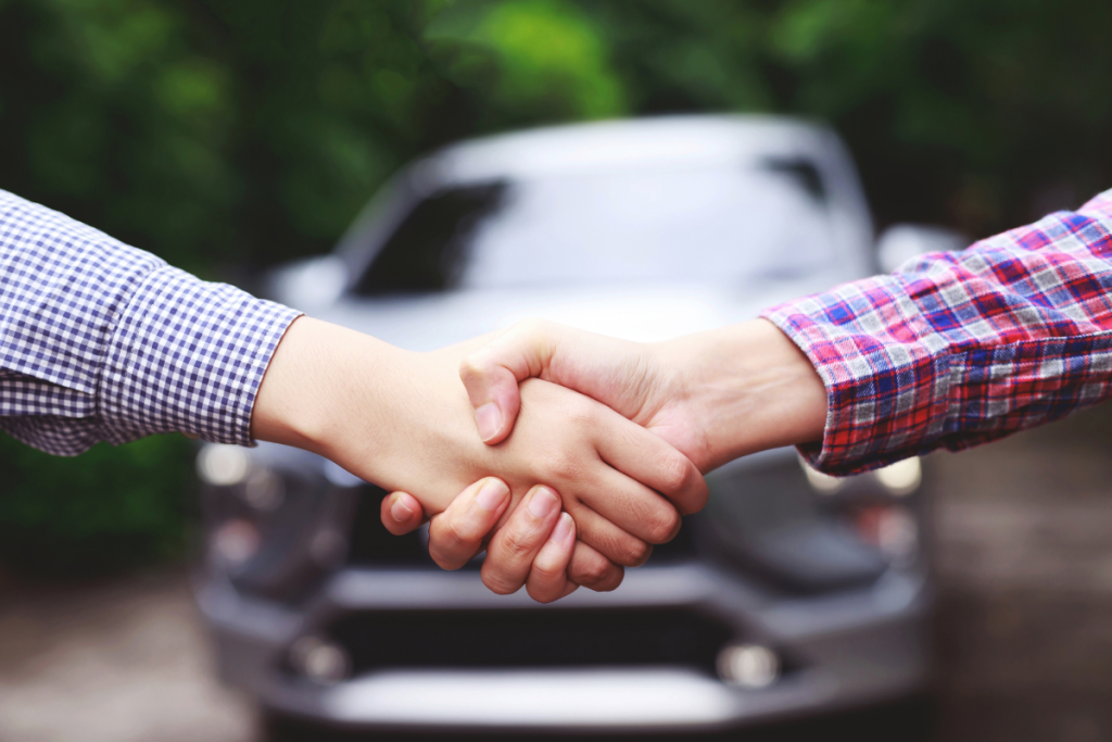 Duas pessoas apertando as mãos com um carro desfocado ao fundo, simbolizando a concretização de um negócio.