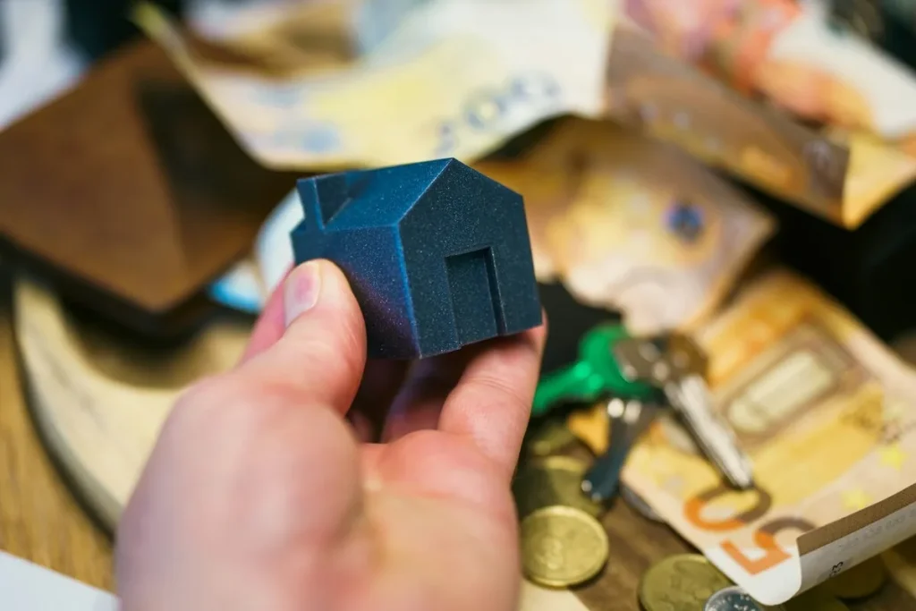 Pessoa segurando uma miniatura de casa com cédulas, moedas e chaves ao fundo, representando aquisição de imóvel via carta de crédito
