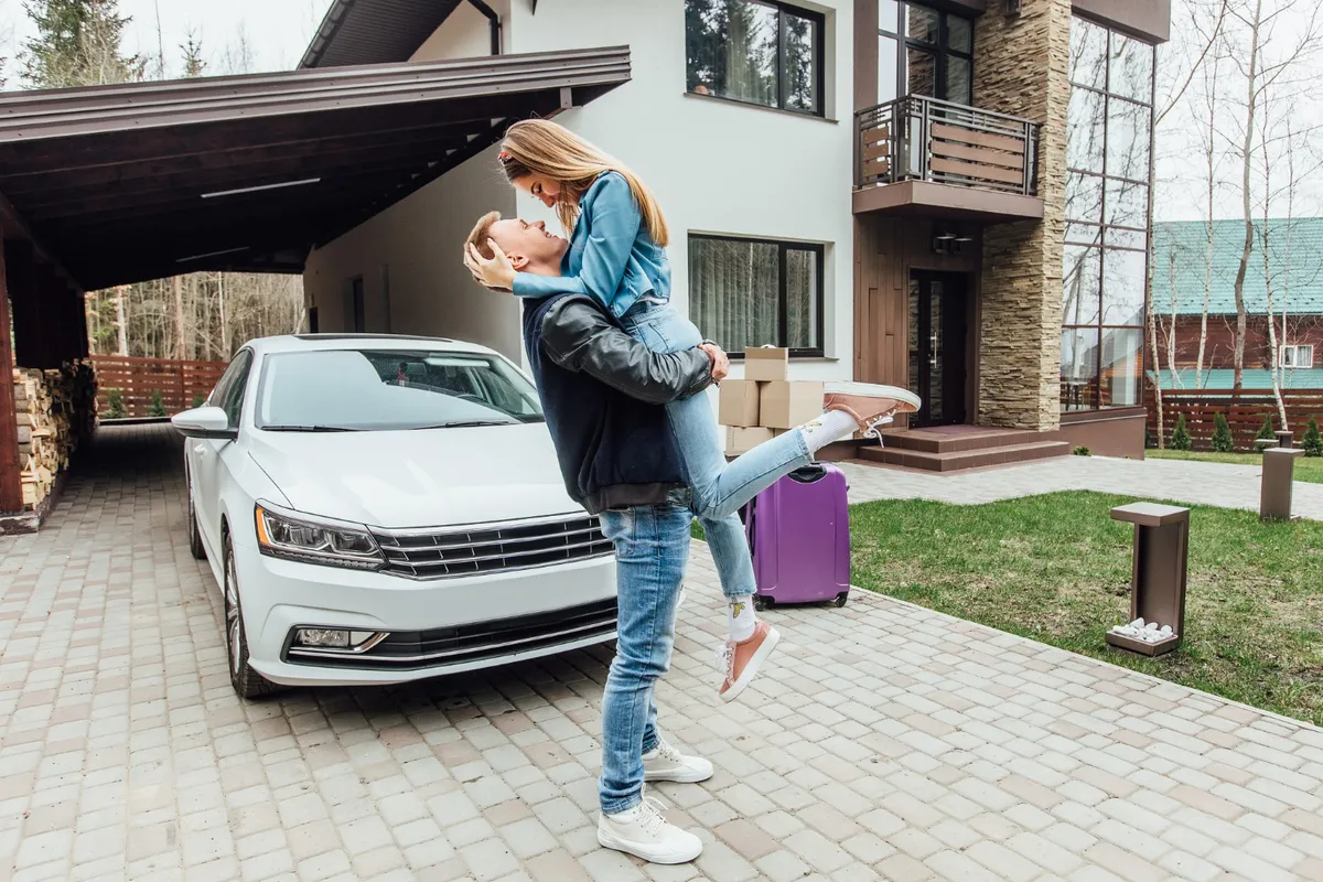 Casal feliz se abraçando em frente a uma casa moderna com carro branco estacionado, malas e caixas de mudança ao fundo.
