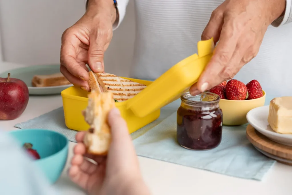 Pessoa colocando sanduíche em pote amarelo de Tupperware ao lado de geleia, morangos e maçã, simbolizando praticidade na alimentação