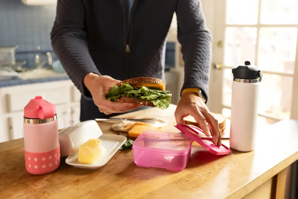 Pessoa preparando sanduíche com folhas verdes para colocar em pote rosa de Tupperware em cozinha iluminada, ao lado de garrafas e manteigueira