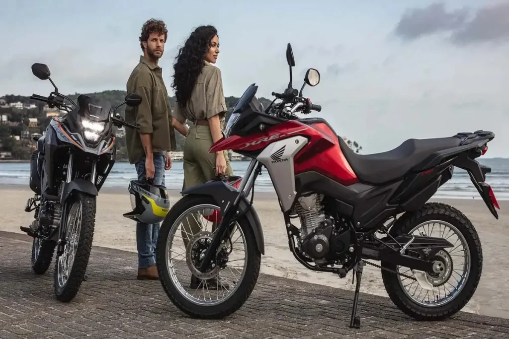 Casal ao lado de duas motos Honda XRE 190 na praia, com destaque para o modelo vermelho, ideal para aventuras e consórcio de motocicleta