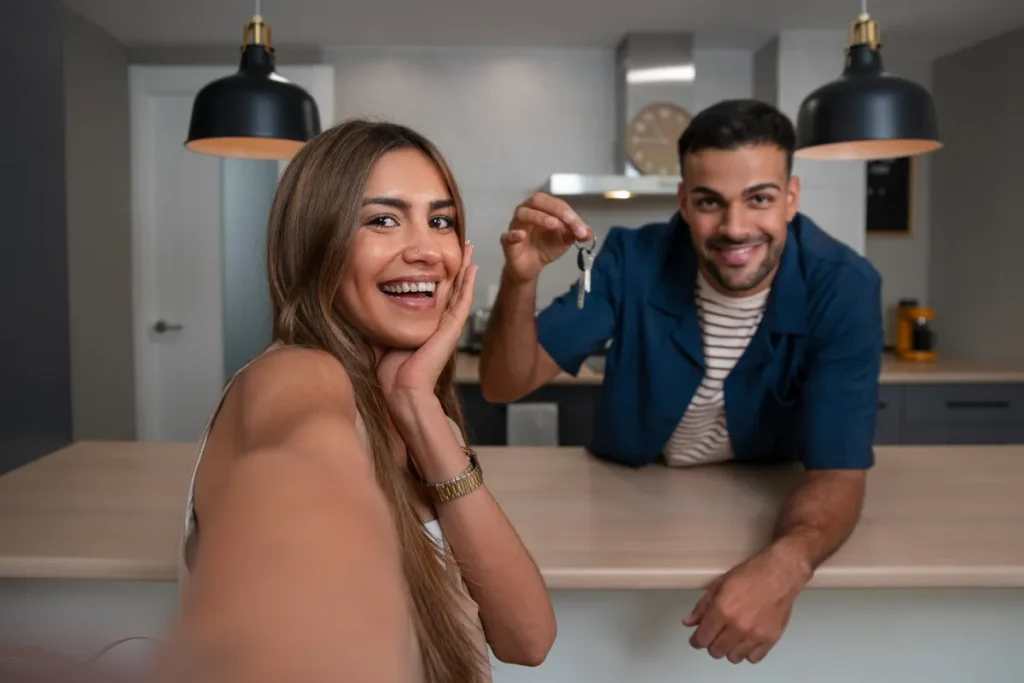 Casal sorridente fazendo selfie na cozinha de casa nova, segurando chaves, representando conquista do imóvel