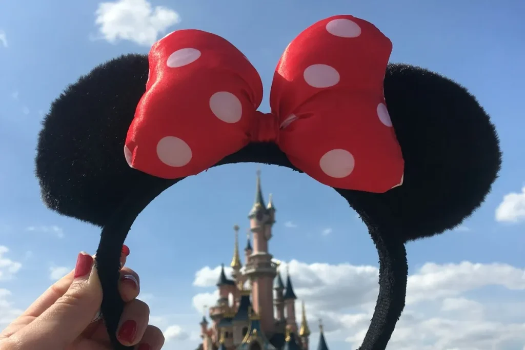 Pessoa segurando uma tiara da Minnie com laço vermelho e orelhas pretas em frente ao castelo da Disney em um dia ensolarado