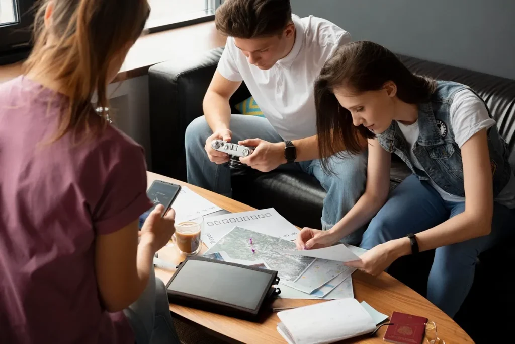 Casal jovem revisando mapas de viagem sobre mesa de madeira, com câmera, smartphone e café, enquanto amiga consulta roteiro em tablet