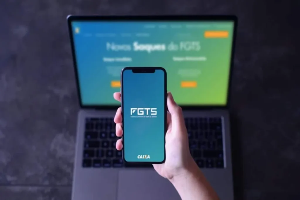 Aplicativo FGTS da Caixa exibido em smartphone com site oficial aberto em notebook ao fundo